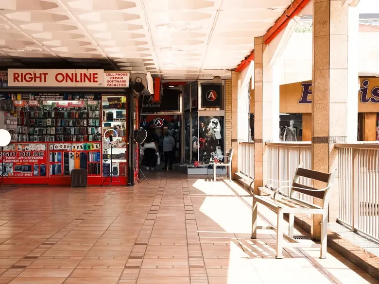 A sunlit covered walkway with shopfronts and a bench